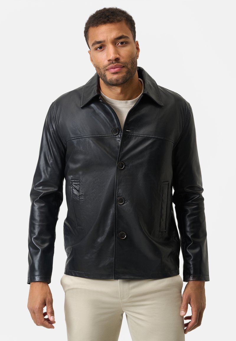Schwarze Lederjacke mit einem Kragen, vorderem Knopfverschluss, zwei Seitentaschen und glatter Textur. Über einem hellen Pullover getragen.