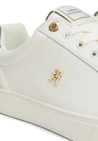 Hvit sneaker med glatt syntetisk materiale, gull logo aksent, runde snorløp og teksturert såle. Minimalistisk design med sømdetaljer.