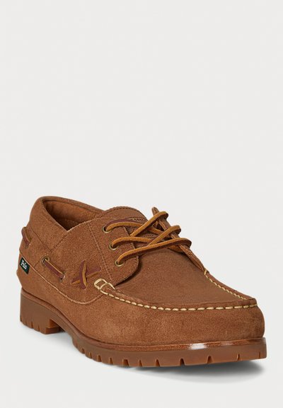 Bruine suède leren bootshoe met veters, crème stiksels en een dikke rubberen zool, getoond vanuit een driekwart vooraanzicht.