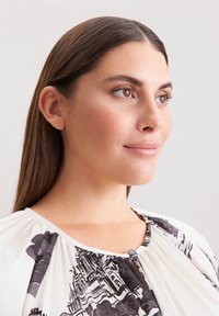 Weiße Bluse mit schwarzem Druckmuster, mit einem gerafften Halsauschnitt und einem Knopfdetail. Die Haare sind gerade und braun und umrahmen das Gesicht.