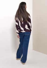 Femme aux longs cheveux bruns portant un pull violet et blanc à motifs et un jean bleu à jambes larges, debout contre un mur bicolore.