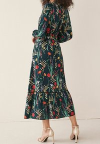 Une robe longue florale vert foncé avec des motifs vibrants rouges, jaunes et violets, dotée d'un ourlet volanté et de manches longues, assortie à des talons dorés.