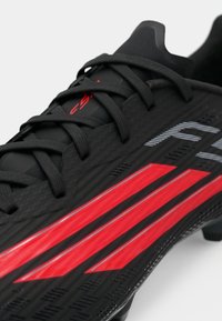 Μαύρα ποδοσφαιρικά παπούτσια με κόκκινες λεπτομέρειες, υφή στην επάνω επιφάνεια, κορδόνια και αεροδυναμικό σχέδιο. Διαθέτει έντονες λωρίδες στο πλάι και σόλα με χιτώνια.