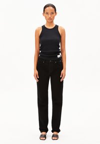 Débardeur noir à côtes associé à un jean noir taille haute. Le modèle se tient droit, portant des sandales à brides ouvertes noires. Design simple et minimaliste.