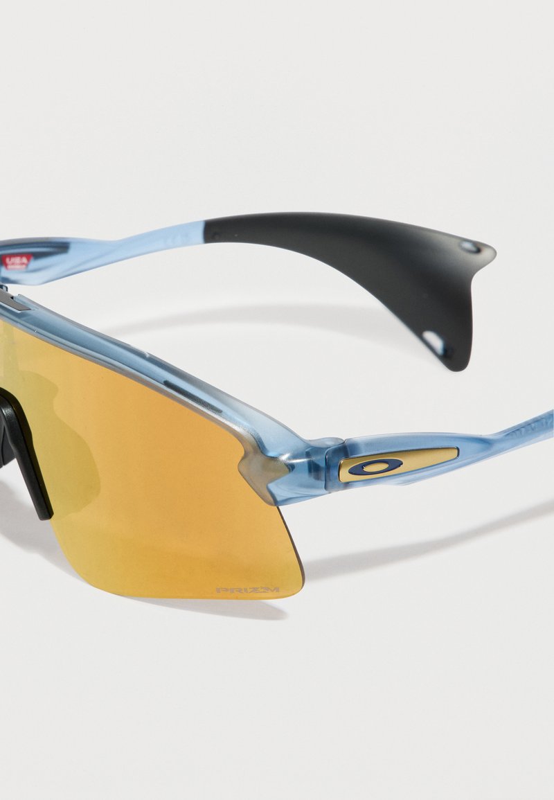 Lunettes de sport semi-cerclées bleues avec verres teintés en jaune et branches en caoutchouc noir sur fond blanc.