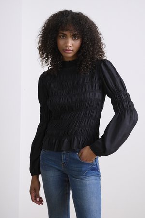 Black smocked blouse with long, puffed sleeves and ruffled hem. Features textured fabric and a high collar, paired with blue jeans.