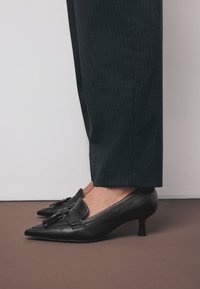 Escarpins noirs en cuir à talons bas avec bouts pointus et un détail décoratif à pompons. Portés avec un pantalon à rayures fines, vue de côté.