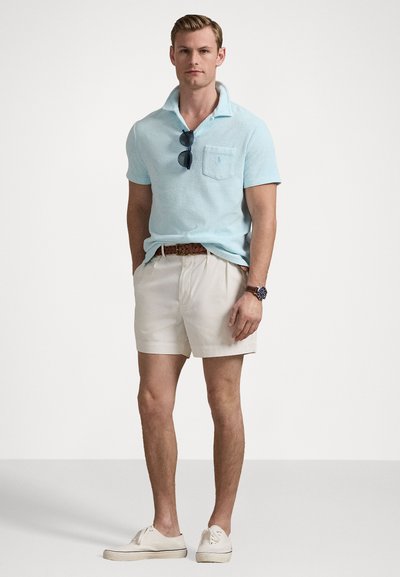 Lichtblauw gestructureerd poloshirt met een borstzak, gedragen met crèmekleurige shorts en witte sneakers. Accessoires zijn onder andere een zonnebril en een polshorloge.