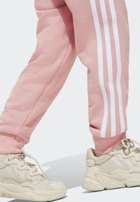 Svetloružové tepláky s bielymi pruhmi po stranách, kombinované s krémovými sieťovanými teniskami s textúrovaným dizajnom a šnúrkami.