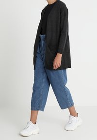 Dark gray cardigan with long sleeves and two front pockets, paired with high-waisted blue denim trousers and white sneakers.