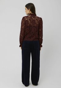 Blouse en dentelle bordeaux avec un col haut et des manches longues, ornée de motifs floraux, associée à un pantalon en velours côtelé bleu marine à jambes larges.