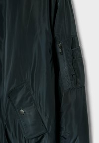 Blouson aviateur vert foncé en tissu lisse et brillant, doté d'une poche zippée sur le bras et d'une grande poche à rabat, présentant un design élégant et une texture épurée.