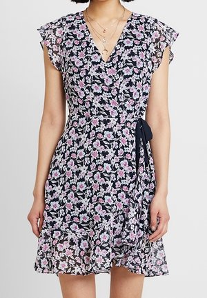 Femme vêtue d'une robe portefeuille noire à imprimé floral rose et blanc, avec des manches courtes volantées et un nœud à la taille, portant des colliers dorés superposés.