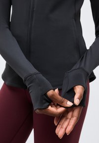 Χέρια που ρυθμίζουν τις τρύπες για τον αντίχειρα σε μαύρο αθλητικό τοπ μακρυμάνικο, που φοριέται με μπορντό κολάν σε άτομο με σκουρόχρωμο δέρμα.