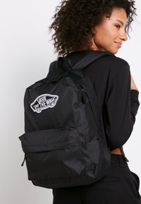 Zaino nero realizzato in tessuto resistente, con un importante patch del logo Vans bianco sul fronte e una tasca con zip sul davanti per la conservazione.