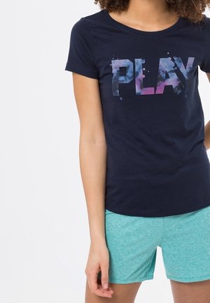 Jeune personne portant un t-shirt bleu marine avec un texte coloré "PLAY" et un short bleu clair, debout devant un fond blanc uni.