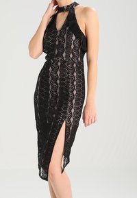Robe en dentelle noire avec encolure halter, présentant une découpe en trou de serrure et une fente sur le côté. Un motif complexe et un tissu texturé mettent en valeur le design.