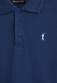 Mörkblå polo-shirt med texturerad väv, tre knappar och en vit golfare-logotyp på vänster bröst. Etiketten visar "POLO CLUB" med storlek "M."