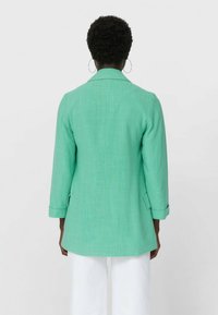 Personne aux cheveux courts et bouclés portant des boucles d'oreilles anneaux, vêtue d'un blazer vert menthe et d'un pantalon blanc, vue de dos.