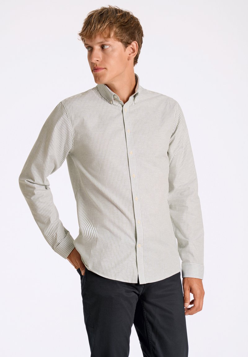 Camisa de botones a rayas en verde claro y blanco, hecha de una tela de textura suave. Presenta un cuello y mangas largas, combinada con pantalones negros.