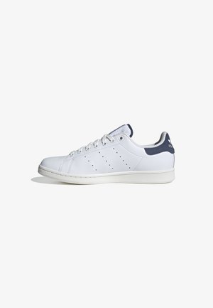 Witte lage sneaker met witte veters, geperforeerd zijpatroon en donkerblauw hielstuk met logo op een witte achtergrond.