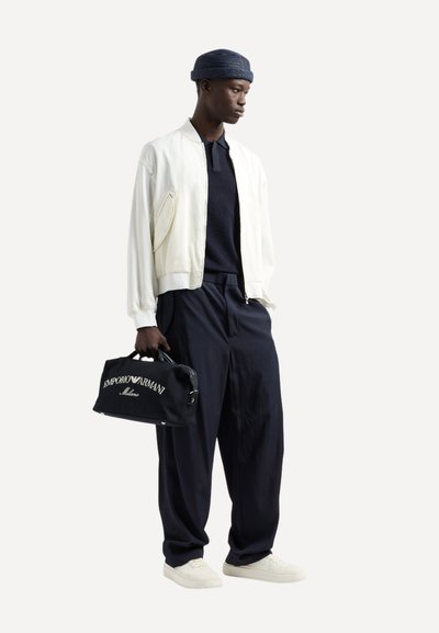 Giacca bomber bianca, polo blu navy, pantaloni blu navy larghi, sneaker bianche e una borsa a mano nera con testo bianco. Un berretto di maglia blu completa il look.