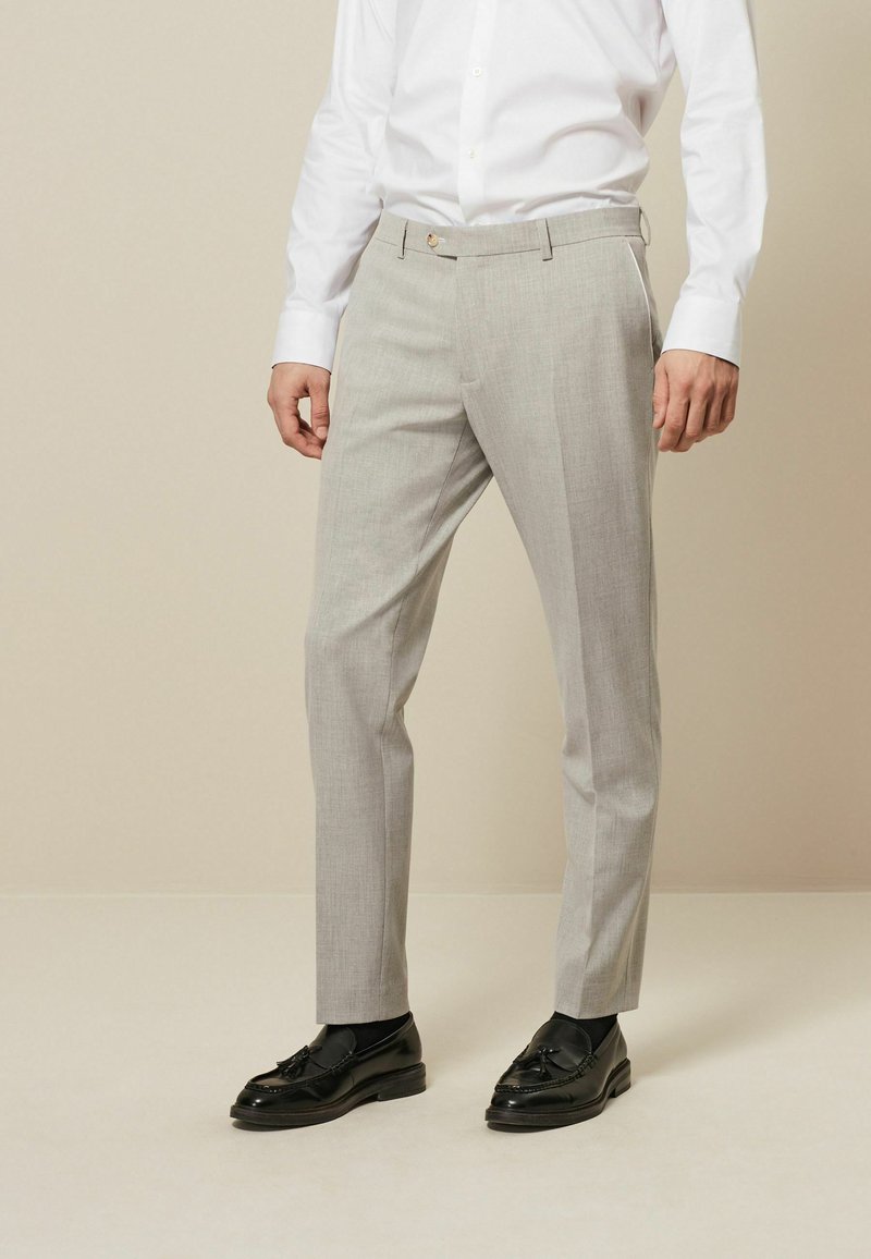 Pantalons tailleur gris clair avec une texture lisse, coupe ajustée, passants de ceinture et fermeture à bouton unique. Associés à des mocassins noirs.