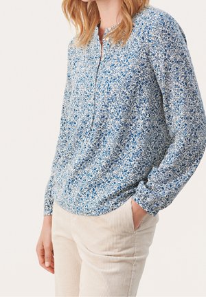 Nainen sinivalkoisessa kukallisessa pitkähihkaisessa paidassa, jossa on osittainen napitus edessä, ja beigeissä cordurohopehkeissä, käsi taskussa.