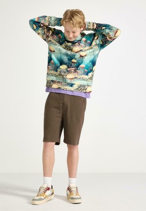 Lyshåret barn, der retter på en farverig sweater med blæksprutte-mønster, iført brune shorts, hvide sokker og flerfarvede sneakers på hvid baggrund.