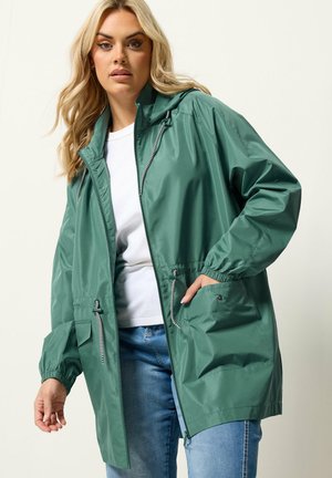 Blonde vrouw die een groene regenjas met capuchon en trekkoorden draagt, een wit shirt en een blauwe spijkerbroek, poserend met één hand in de zak.