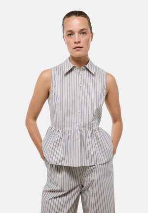 Femme aux cheveux attachés porte un haut sans manches à col boutonné et un pantalon rayé assorti, les mains dans les poches, sur fond blanc.