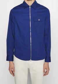Camisa azul de manga larga con cremallera en la parte delantera, cuello puntiagudo, un bolsillo en el pecho y detalles de puños con botón. Fabricada con una tela suave.