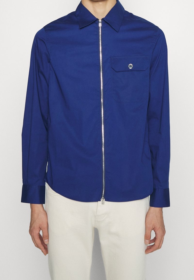 Camisa azul de manga larga con cremallera en la parte delantera, cuello puntiagudo, un bolsillo en el pecho y detalles de puños con botón. Fabricada con una tela suave.