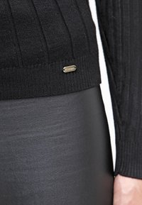 Pull noir côtelé avec une étiquette logo dorée à l'ourlet, associé à un pantalon noir élégant. Accent sur la texture et les matériaux contrastés.