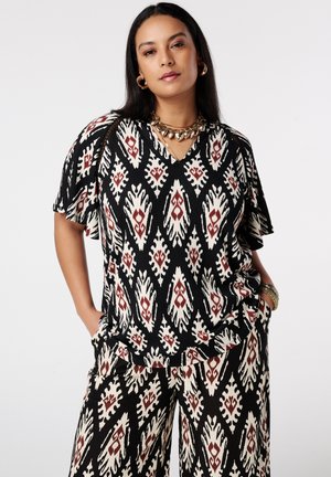 Femme portant un ensemble assorti noir et blanc à motifs avec des bijoux en or, les mains dans les poches, debout devant un fond uni.