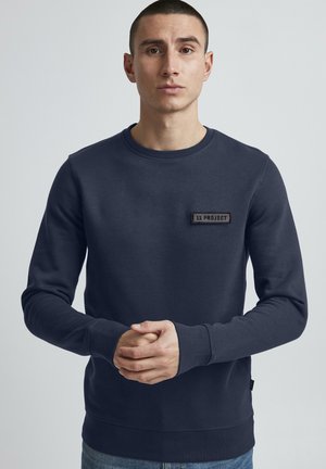 Marineblå sweatshirt med rund hals og lange ærmer. Har en struktureret sort lap med teksten "11 PROJECT" på forsiden.