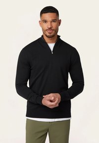 Zwarte half-zip trui met lange mouwen, gemaakt van een glad gebreid stof, met een geribbelde zoom en manchetten, gedragen over een wit shirt.