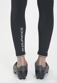 Schwarze Sportleggings mit einem silbernen "ENDURANCE"-Logo, die ein enganliegendes Design und graue Akzente an den Bündchen aufweisen, kombiniert mit schwarzen Schuhen.