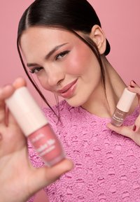 Dos botellas de cosméticos con tapas de color rosa claro, una de ellas etiquetada como "Wonder Me", sostenidas por una persona con una prenda texturizada rosa, sobre un fondo rosa.