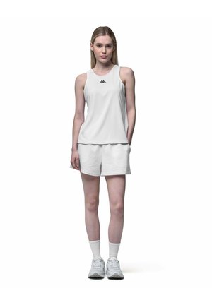 Jeune femme portant un débardeur de sport blanc, un short assorti, des chaussettes blanches et des baskets, debout devant un fond blanc uni.