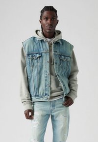 Šviesiai mėlynas džinsinis liemenėlis su išeidžiomis vietomis ir sidabrinės spalvos detalėmis, dėvimas ant pilkos spalvos džemperio su raišteliu. Šviesiai plauti, nudėvėti džinsai.