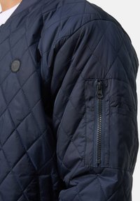 Marineblå quilted bomberjakke med et rundt logo på brystet og en lynlås lomme på ærmet. Glat, polstret tekstur for varme.