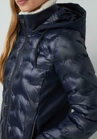 Mornarska puf jakna z bleščečo površino, ki vključuje kapuco, zadrgo in sprednje žepke. Oblikovanje vsebuje šivane, prešite dele.