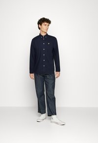 Camisa azul marinho de manga longa com botões, apresentando um pequeno logótipo bordado no peito, combinada com calças de ganga cinzas e ténis brancos.