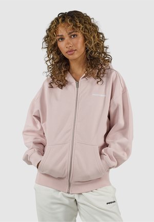 Femme aux cheveux bouclés portant un sweat à capuche rose clair zippé et un pantalon blanc avec des logos de marque, debout les mains dans les poches du sweat.