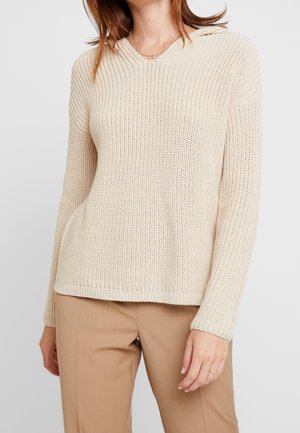 Woman wearing cream-colored ribbed knit sweater with long sleeves and beige pants, standing against light background.