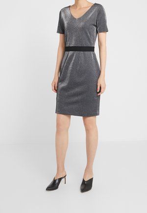 Kobieta w srebrnej błyszczącej sukience do kolan z krótkim rękawem, dekoltem w serek i czarnym paskiem w talii, w połączeniu z czarnymi spiczastymi czółenkami mule.