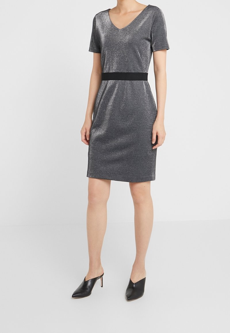 Femme portant une robe pailletée argentée mi-genoux à manches courtes, décolleté en V et ceinture noire, associée à des mules noires pointues.