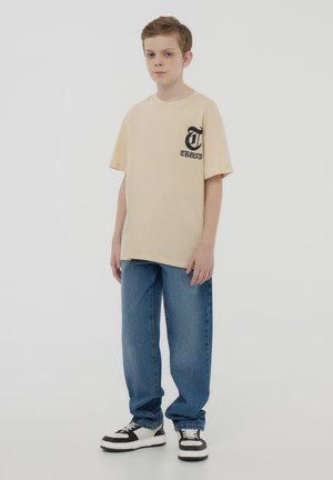 T-shirt di cotone beige con logo nero sul lato sinistro del petto, abbinata a jeans di denim blu e sneaker bianche e nere.