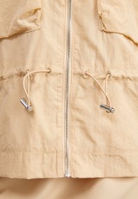 Ljust beige jacka med dragkedja, justerbara dragsko och silverfärgade detaljer. Tillverkad av ett lätt, något strukturerat tyg.